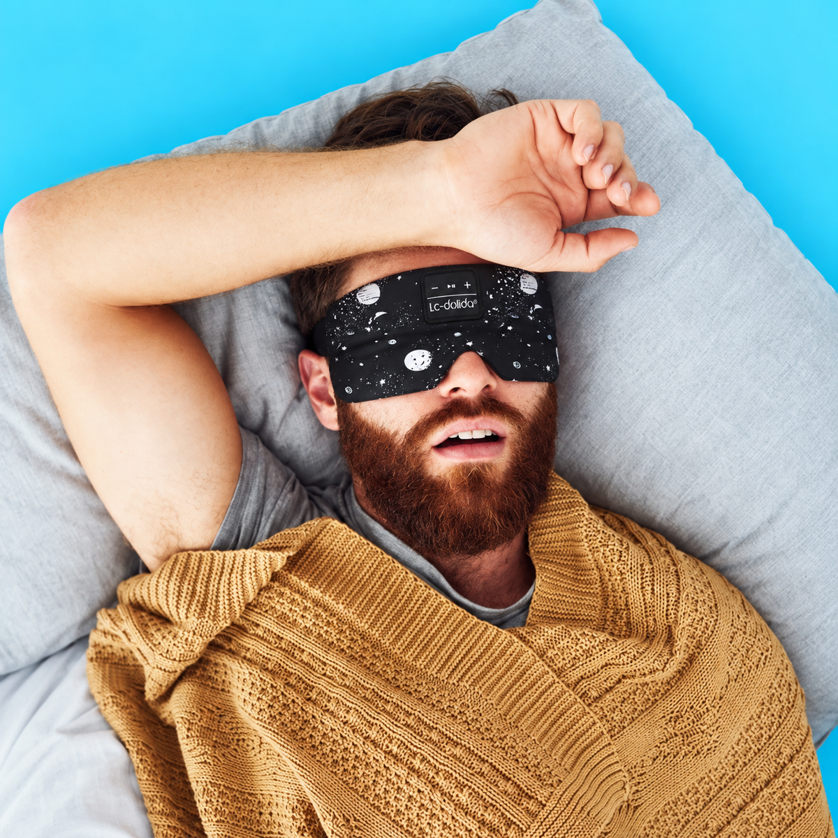 Man resting in bed wearing a black space-print Bluetooth sleep mask